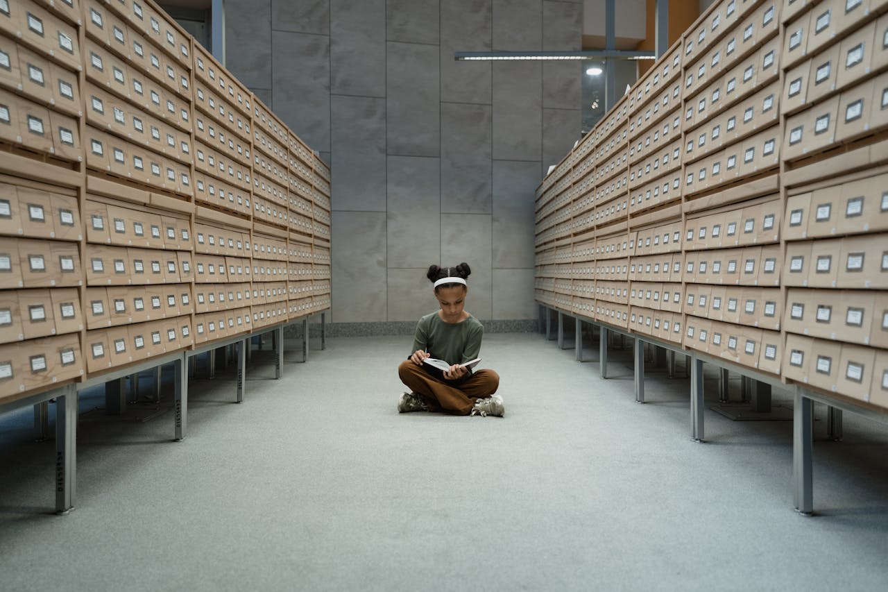 A Girl Sitting on the Floor Reading a Book Between Wooden Drawers